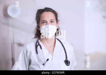 Jeune femme positive médecin en uniforme blanc et port de stéthoscope masque médical de protection debout dans un bureau médical moderne et regardant à l'appareil photo Banque D'Images