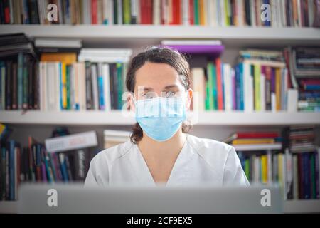 Jeune femme médecin sérieux portant un uniforme blanc et un masque médical travailler sur un ordinateur portable avec des gants en latex assis au bureau clinique moderne Banque D'Images
