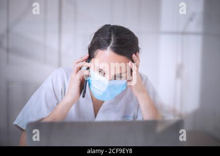 Jeune femme médecin sérieux portant un uniforme blanc et un masque médical travailler sur un ordinateur portable avec des gants en latex assis au bureau clinique moderne Banque D'Images