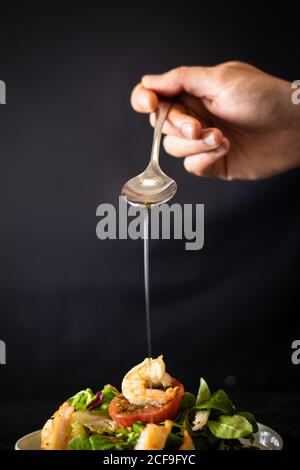 cuisson non reconnaissable ajoutant une cuillerée d'huile à une salade de légumes délicieuse avec crevettes sur fond noir Banque D'Images