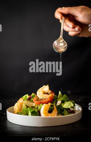 cuisson non reconnaissable ajoutant une cuillerée d'huile à une salade de légumes délicieuse avec crevettes sur fond noir Banque D'Images
