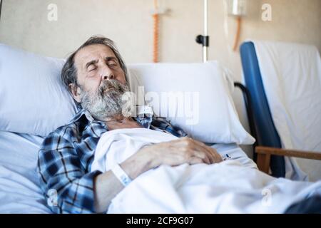 Homme calme et âgé avec une barbe sous une couverture sur le lit dans la salle d'hôpital et de dormir Banque D'Images