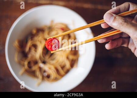 Du dessus du foyer doux de la personne de récolte tenant la tomate de cerise par des baguettes en bois tout en appréciant les nouilles sur fond flou Banque D'Images
