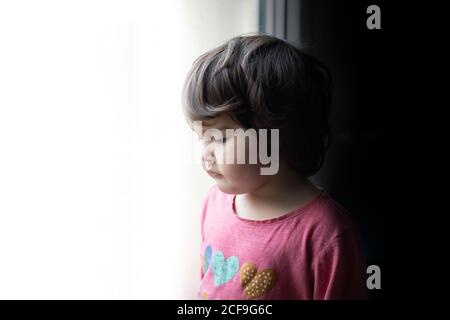 Vue latérale de la triste petite fille debout près de la fenêtre et regarder tout en passant du temps à la maison Banque D'Images