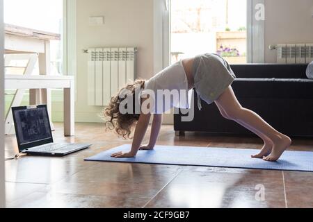 Vue latérale de la petite fille faisant face à l'asana de chien tout en pratiquant le yoga devant un ordinateur portable dans le salon à la maison Banque D'Images