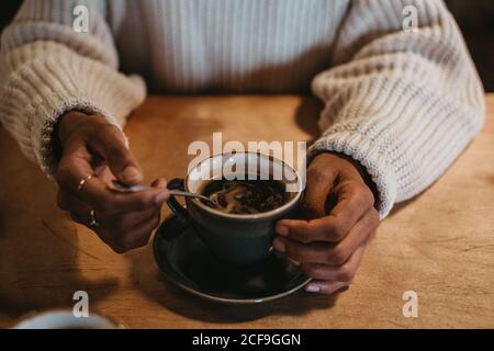De dessus, crop mains de femelle méconnaissable assis au bois table contenant du café chaud dans une tasse rustique Banque D'Images