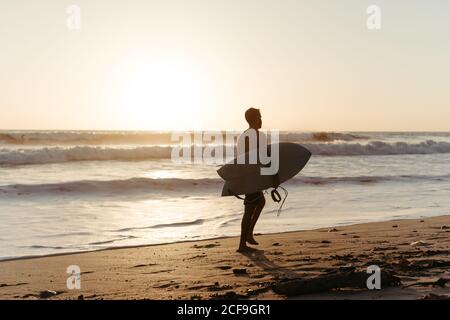 Vue arrière de la silhouette d'homme anonyme tenant une planche de surf pendant la marche le long de la plage de sable en été pendant le coucher du soleil Banque D'Images