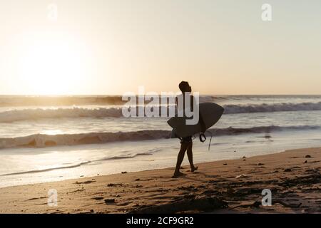 Vue arrière de la silhouette d'homme anonyme tenant une planche de surf pendant la marche le long de la plage de sable en été pendant le coucher du soleil Banque D'Images