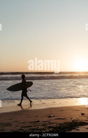 Vue latérale d'un homme anonyme tenant une planche de surf pendant la marche le long de la plage de sable en été pendant le coucher du soleil Banque D'Images