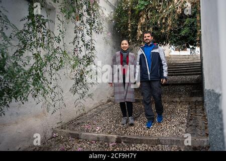 Contenu multi-ethnique couple en vacances dans la tenue de vêtements décontractés mains tout en marchant sur une vieille allée rockée avec un vert exotique Plantes à Albaicin à Grenade Banque D'Images