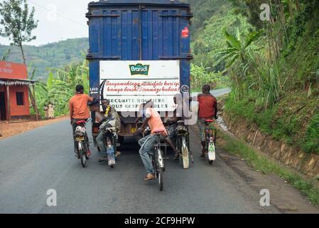 Vue arrière du groupe de jeunes hommes de la région qui font du vélo et suivre le camion-conteneur avec l'emblème de la compagnie de brasserie sur Route rurale parmi les forêts au Burundi en Afrique Banque D'Images