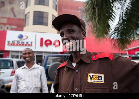 Garde de sécurité masculine gaie portant un uniforme marron avec l'emblème de compagnie de sécurité privée regardant la caméra tout en se tenant contre local Faites des emplettes au Burundi avec un homme souriant flou en arrière-plan Banque D'Images