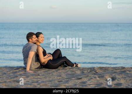 Vue latérale d'un jeune couple multiracial joyeux dans un salon sportswear sur une plage de sable, en vous reposant après un entraînement et en profitant du temps ensemble Banque D'Images