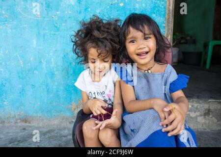 Dili, Timor oriental - 09 AOÛT 2018: Petite fille souriante regardant la caméra et sœur plus jeune jouant élastique tout en étant assis ensemble à côté de la maison pauvre Banque D'Images