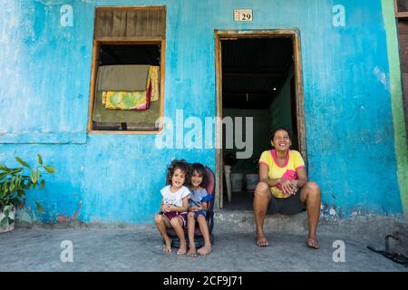 Dili, Timor oriental - 09 AOÛT 2018: Femme gaie et petites filles regardant la caméra tout en étant assis à la porte de la maison pauvre Banque D'Images