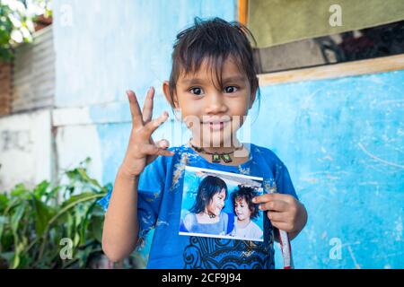Dili, Timor oriental - 09 AOÛT 2018 : petite fille gaie qui chante et regarde l'appareil photo tout en se tenant à côté de la maison pauvre avec une photo à la main heureuse avec le cadeau d'un photographe professionnel Banque D'Images
