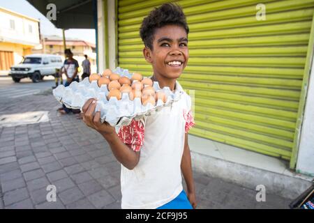 Dili, Timor oriental - 09 AOÛT 2018 : garçon asiatique pauvre et gai marchant dans la rue de la ville et portant un plateau en carton avec des œufs Banque D'Images