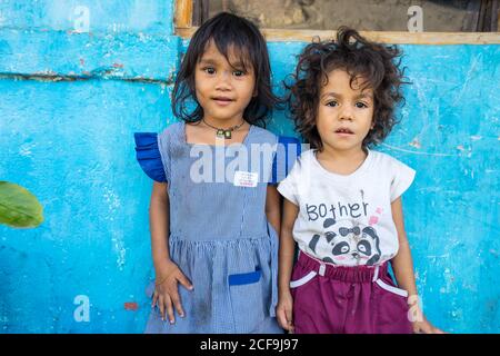 Dili, Timor oriental - 09 AOÛT 2018: Petite fille souriante regardant la caméra et sœur plus jeune jouant élastique tout en se tenant ensemble à côté de la maison pauvre Banque D'Images
