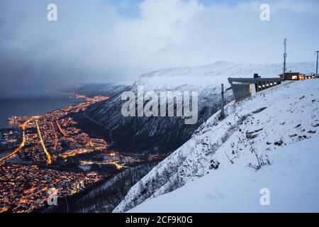 Paysage incroyable de la ville avec lumières dorées situées le long de la côte dans la vallée au pied de collines enneigées sous ciel nuageux au crépuscule Banque D'Images