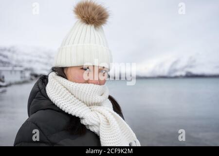 Jeune femme avec une veste noire et un chapeau chaud blanc et une écharpe regardant l'appareil photo sur fond gris flou en Norvège Banque D'Images