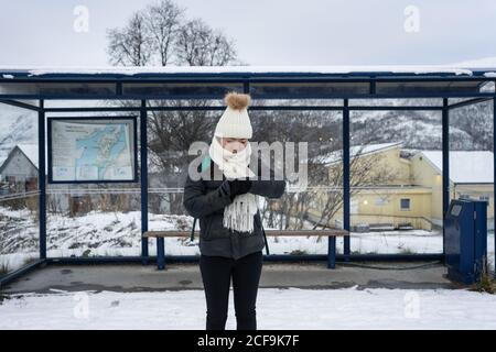 Une jeune femme asiatique vêtue de vêtements chauds regardant sa montre en attendant son transport à un arrêt de bus par une route enneigée en Norvège Banque D'Images