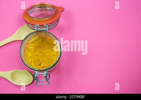 Vue de dessus sur un pot de conservation isolé en verre avec poudre curcuma et deux cuillères à bois, fond rose. Espace pour le texte. (accent sur la poudre) Banque D'Images