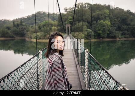 Vue latérale d'une femme asiatique rêveuse en tenue décontractée avec caméra photo debout sur le pont suspendu au-dessus du lac à proximité vaste forêt et regarder l'appareil photo Banque D'Images