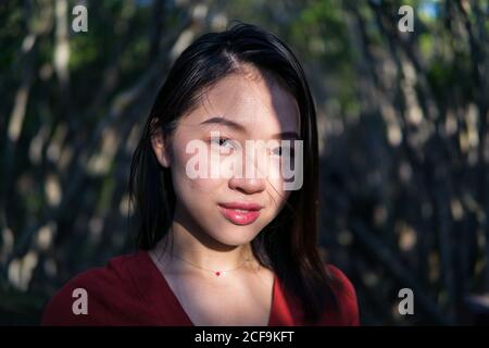 Jeune femme asiatique réfléchie en tenue décontractée et petit collier regarder la caméra en forêt à la lumière du jour Banque D'Images