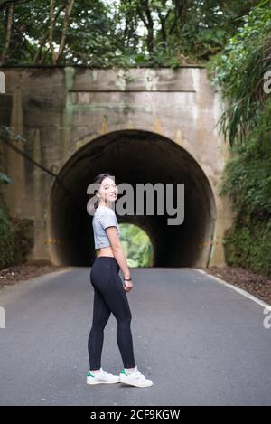 Vue latérale de la jeune femme asiatique élancée dans des leggings debout sur la route asphaltée devant le tunnel, près du mur couvert avec des plantes vertes et regarder la caméra Banque D'Images