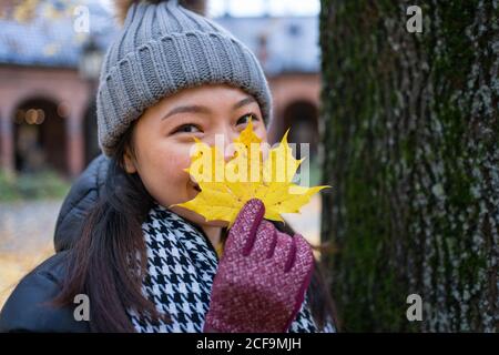 Heureuse jeune femme asiatique ethnique vêtue de vêtements chauds, regardant l'appareil photo et couvrant le visage avec une feuille d'érable jaune pendant qu'elle se tient à la cathédrale d'Oslo en Norvège Banque D'Images