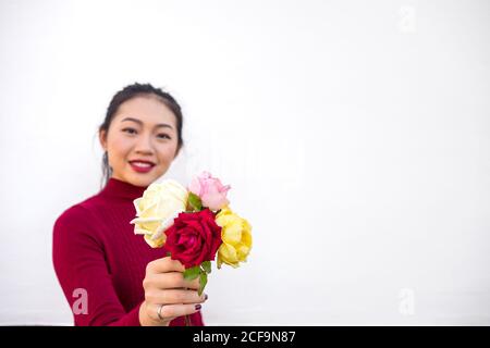 Jeune femme asiatique en turtleneck bordeaux tenant et présentant un bouquet de roses colorées tout en regardant l'appareil photo avec le sourire Banque D'Images