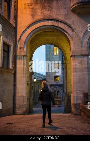 Vue arrière de la femme en veste chaude et chapeau marchant le long de la rue sous l'arche dans la lumière du soir à Saint-Marin, Italie Banque D'Images