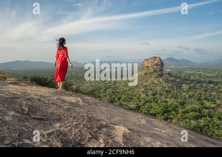 Vue arrière de la femme méconnue en robe rouge bénéficiant d'une vue imprenable sur la forteresse de roche solitaire Sigiriya au milieu du plateau avec forêt tropicale dense et verte de la montagne Pidurangala sous le ciel bleu au Sri Lanka Banque D'Images