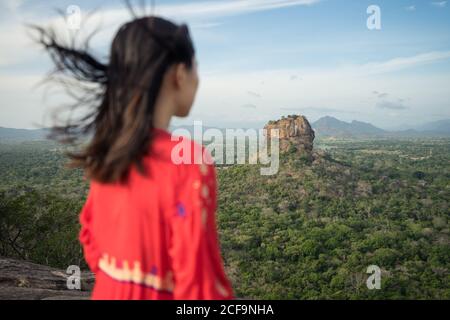 Vue arrière de la femme méconnue en robe rouge bénéficiant d'une vue imprenable sur la forteresse de roche solitaire Sigiriya au milieu du plateau avec forêt tropicale dense et verte de la montagne Pidurangala sous le ciel bleu au Sri Lanka Banque D'Images