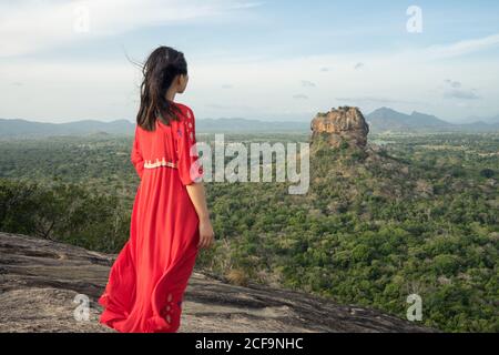 Vue arrière de la femme méconnue en robe rouge bénéficiant d'une vue imprenable sur la forteresse de roche solitaire Sigiriya au milieu du plateau avec forêt tropicale dense et verte de la montagne Pidurangala sous le ciel bleu au Sri Lanka Banque D'Images