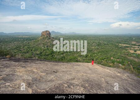 Vue arrière de la femme méconnue en robe rouge bénéficiant d'une vue imprenable sur la forteresse de roche solitaire Sigiriya au milieu du plateau avec forêt tropicale dense et verte de la montagne Pidurangala sous le ciel bleu au Sri Lanka Banque D'Images