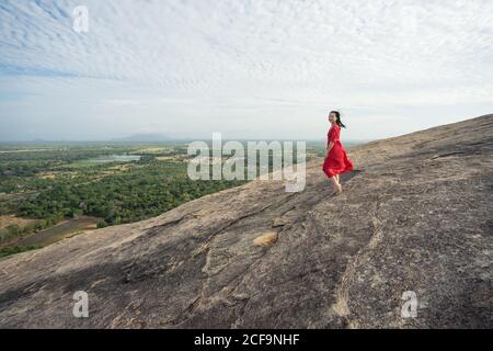 Vue latérale de la femme en robe rouge appréciant la vue étonnante de la forteresse de roche solitaire Sigiriya au milieu du plateau avec forêt tropicale dense vert de la montagne Pidurangala sous le ciel bleu au Sri Lanka Banque D'Images