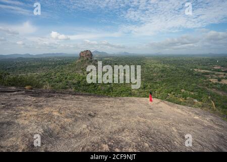 Vue latérale d'une femme méconnaissable en robe rouge bénéficiant d'une vue imprenable sur la forteresse de roche solitaire Sigiriya au milieu d'un plateau avec une forêt tropicale dense et verte depuis la montagne Pidurangala sous le ciel bleu du Sri Lanka Banque D'Images