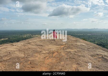 Vue arrière de la femme méconnue en robe rouge bénéficiant d'une vue imprenable sur la forteresse de roche solitaire Sigiriya au milieu du plateau avec forêt tropicale dense et verte de la montagne Pidurangala sous le ciel bleu au Sri Lanka Banque D'Images