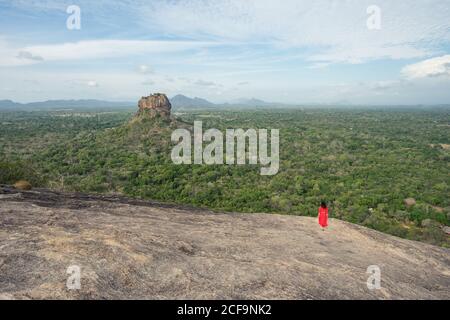 Vue arrière de la femme méconnue en robe rouge bénéficiant d'une vue imprenable sur la forteresse de roche solitaire Sigiriya au milieu du plateau avec forêt tropicale dense et verte de la montagne Pidurangala sous le ciel bleu au Sri Lanka Banque D'Images