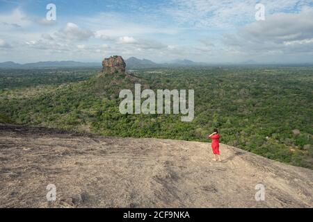 Vue arrière de la femme méconnue en robe rouge bénéficiant d'une vue imprenable sur la forteresse de roche solitaire Sigiriya au milieu du plateau avec forêt tropicale dense et verte de la montagne Pidurangala sous le ciel bleu au Sri Lanka Banque D'Images