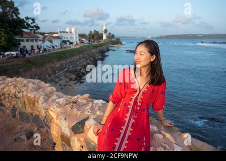 Femme asiatique satisfaite en vacances dans une robe lumineuse colorée assise sur la clôture rockée du quai tropical et en regardant loin Galle au Sri Lanka Banque D'Images