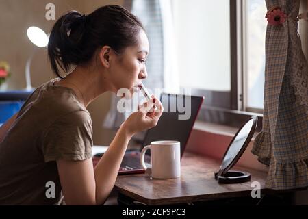 Vue latérale d'une jeune femme ethnique sérieuse en vêtement décontracté s'asseoir à la table devant le miroir et appliquer le rouge à lèvres Banque D'Images
