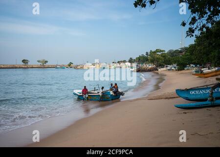 Tangalle, Sri Lanka - 28 juillet 2019 : pêcheurs ethniques en vêtements décontractés, le bateau bleu sur le rivage sablonneux vide Banque D'Images