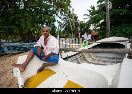 Tangalle, Sri Lanka - 28 juillet 2019 : pêcheur ethnique senior en vêtements légers décontractés assis sur le bateau et regardant la caméra Banque D'Images