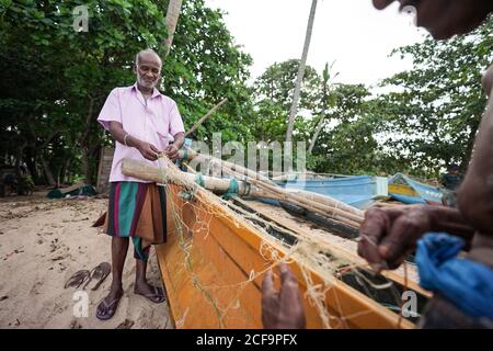 Tangalle, Sri Lanka - 28 juillet 2019: Pêcheur ethnique en vêtements traditionnels décontractés debout avec des armes akimbo et regardant vers le bas près du bateau avec le crâne à la plage de sable Banque D'Images