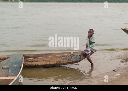Ouganda - novembre, 26 2016: Vue latérale d'un garçon africain dans des vêtements sales et décontractés assis dans un bateau de pêche en chiche près de la côte de la rivière Banque D'Images