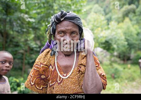 Ouganda - novembre, 26 2016: Femme africaine senior de corps entier regardant la caméra et le sac de transport sur le dos tout en marchant sur le chemin dans la forêt Banque D'Images