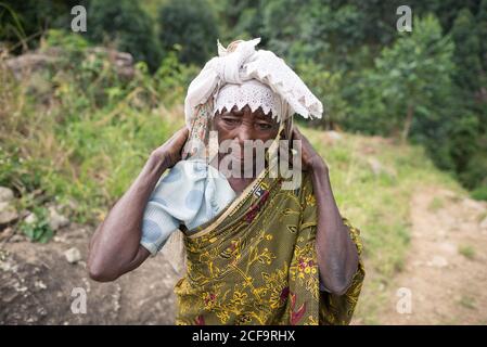 Ouganda - novembre, 26 2016: Femme africaine senior de corps entier regardant loin et portant sac sur le dos tout en marchant sur le chemin dans la forêt Banque D'Images