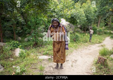 Ouganda - novembre, 26 2016: Femme africaine senior de corps entier regardant la caméra et le sac de transport sur le dos tout en marchant sur le chemin dans la forêt Banque D'Images